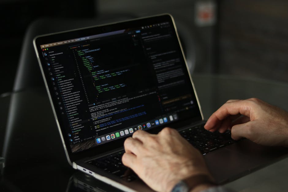 A person working on a laptop displaying a dark-themed coding interface with lines of colorful code, resting on a dark surface. The individual’s hands are active on the keyboard, positioned in a natu