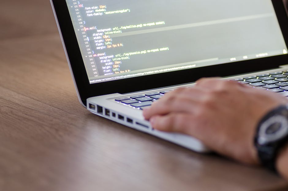 A person working on a laptop displaying a dark-themed coding interface with lines of colorful code, resting on a dark surface. The individual’s hands are active on the keyboard, positioned in a natu