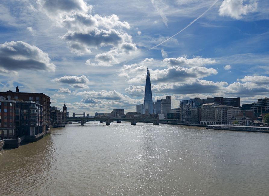 A wide view of the London Bridge Riverfront taken during daytime, showcasing a calm waterway with reflections of the partly cloudy sky overhead. Modern buildings and residential apartments line the ri